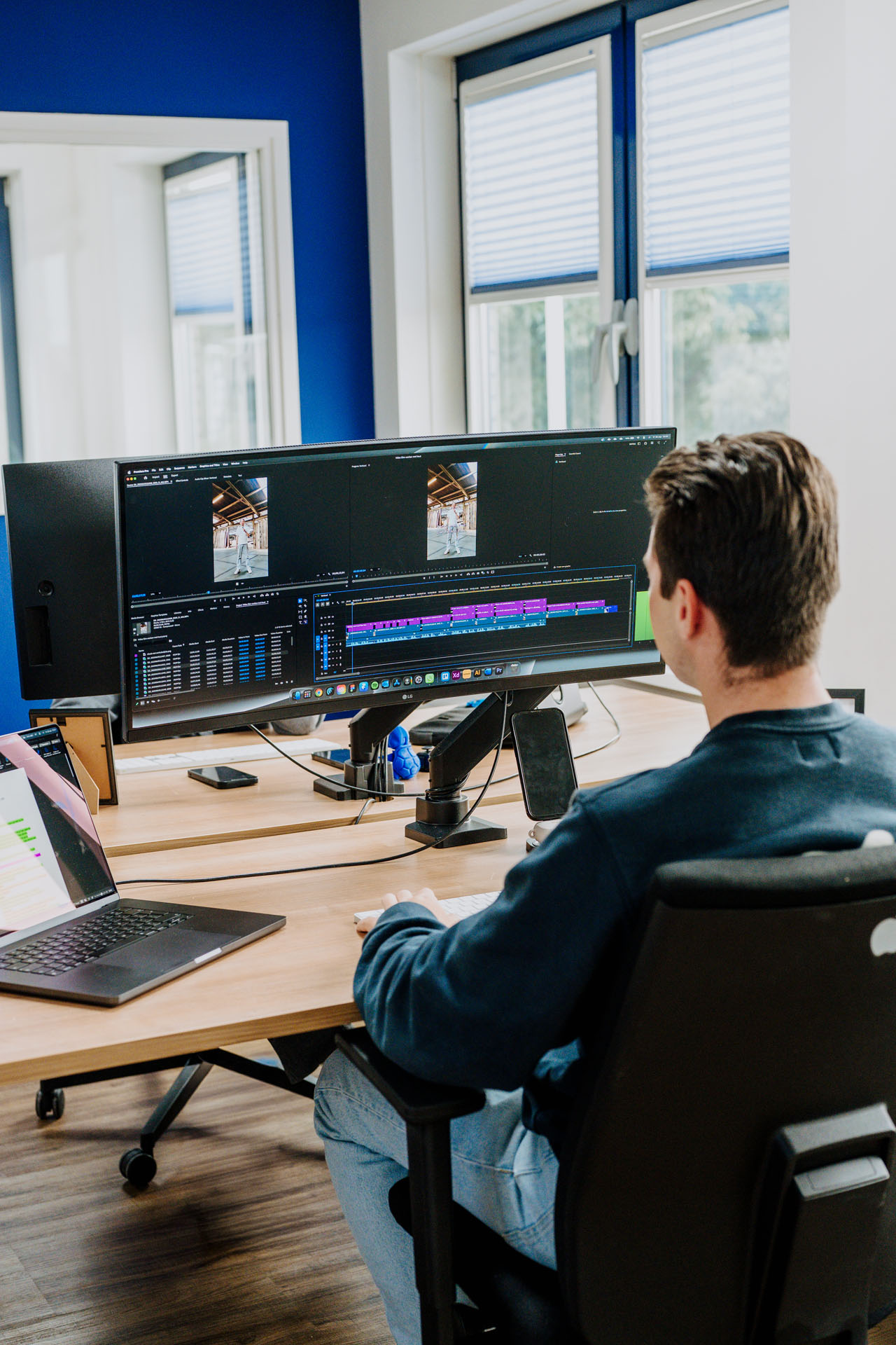 Man zit achter een bureau en werkt aan videobewerking op een groot ultrabreed beeldscherm in een modern kantoor met blauwe muren en grote ramen.