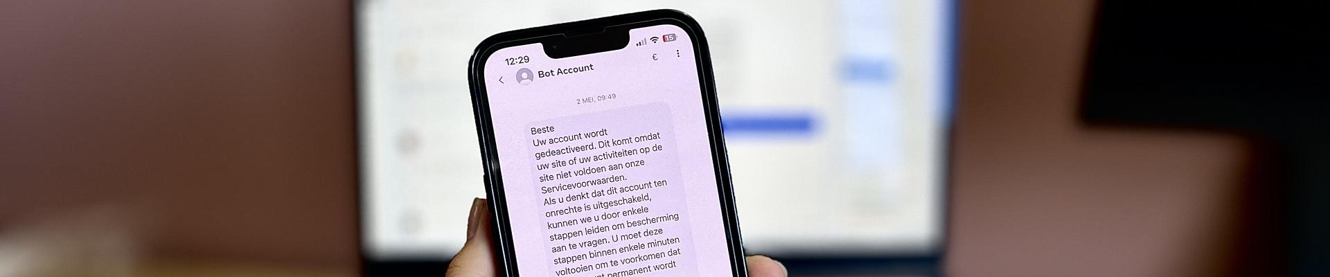 Hand die telefoon vasthoudt met daarop een bericht van Bot Account, op de achterkant geblurd een computer