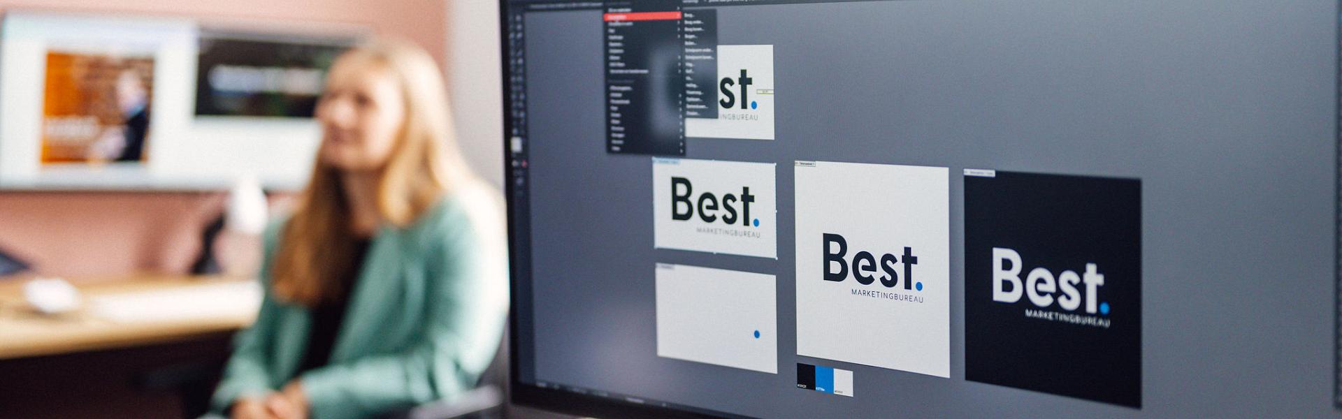 Scherm met verschillende vormen van het Best logo er op. Vrouw aan bureau vanaf de zijkant op de achtergrond.