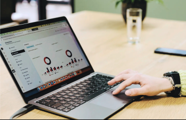 Laptop op houten tafel toont data en hand op toetsenbord klikt iets aan in kantooromgeving.