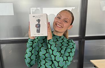 Vrouw glimlacht en houdt een doos met DJI Osmo Pocket 3 Creator Combo camera omhoog in een kantoorruimte.