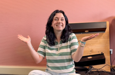 Vrouw met donker haar zit lachend op de grond met haar armen omhoog, voor een roze muur en kabels op de achtergrond