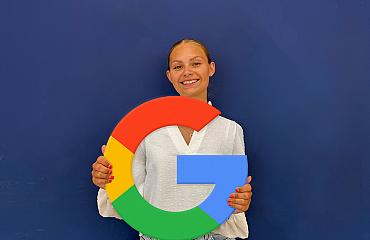 Vrouw die glimlachend een groot Google-logo vasthoudt, gekleed in een witte blouse en lichtblauwe jeans, voor een effen blauwe achtergrond.