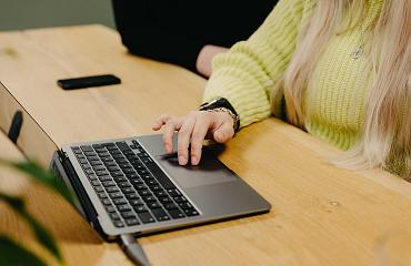 Close-up van een persoon met lang blond haar en een groene trui die aan het werk is op een laptop aan een houten tafel.