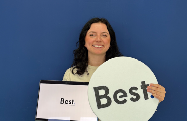 Een vrouw staat lachend voor een blauwe muur terwijl ze een laptop en een bord met het logo “Best.” vasthoudt.