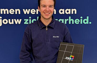 Jonge man met donkerblond haar en een blauwe trui poseert lachend voor een blauwe wand met de tekst “Samen werken aan jouw zekerheid.” Hij houdt een zwarte map met het PWA-logo vast.