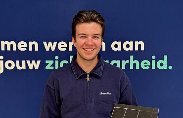 Jonge man met donkerblond haar en een blauwe trui poseert lachend voor een blauwe wand met de tekst “Samen werken aan jouw zekerheid.” Hij houdt een zwarte map met het PWA-logo vast.