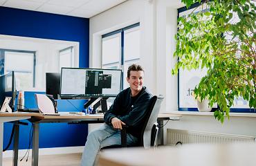 Man die glimlachend achter zijn bureau zit in een modern kantoor met blauwe muren, meerdere beeldschermen en een grote groene plant bij het raam.