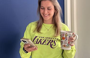 Vrouw die glimlachend op haar telefoon kijkt terwijl ze een mok vasthoudt, gekleed in een felgele Los Angeles Lakers-trui, voor een blauwe muur.