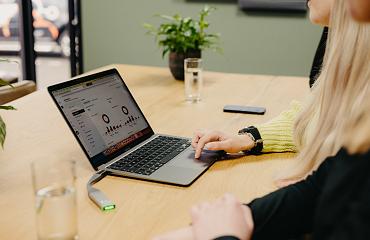 Twee mensen die samenwerken achter een laptop waarop grafieken en statistieken te zien zijn, aan een houten tafel met glazen water en planten.