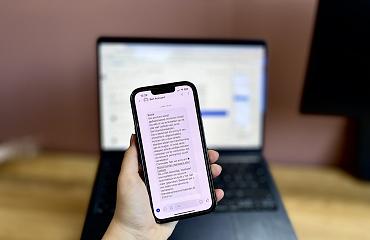Hand die telefoon vasthoudt met daarop een bericht van Bot Account, op de achterkant geblurd een computer