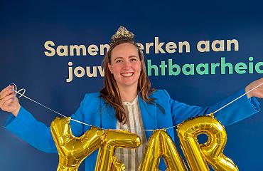 Medewerkster staat lachend voor een blauwe wand en houdt gouden ballonnen met het woord “YEAR” vast, als symbool voor een jubileum of viering.