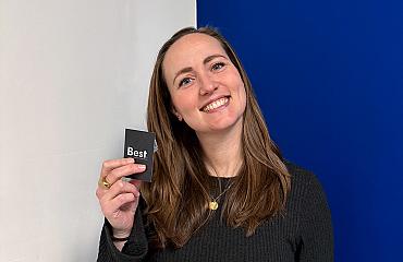 Vrouw met lang bruin haar houdt een zwart visitekaartje met het logo Best vast en glimlacht tegen een witte en blauwe achtergrond.