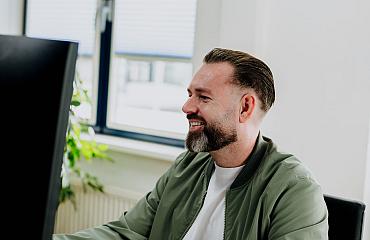 Man met baard lachend achter een computerscherm met een kopje koffie