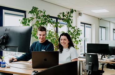 Twee collega’s werken samen achter een bureau met laptop en monitor in een licht kantoor met grote ramen en groene planten op de achtergrond.