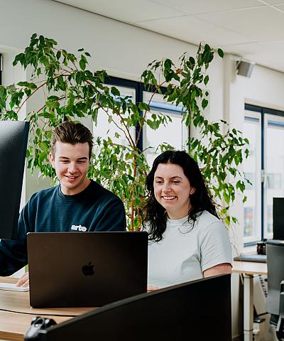 Twee collega’s werken samen achter een bureau in een modern kantoor met veel licht en groene planten.