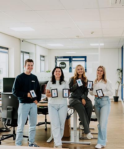 Vier collega’s poseren lachend in een modern kantoor, elk met een tablet in hun handen waarop een illustratie van henzelf te zien is.