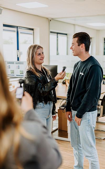 Vrouw houdt een microfoon vast terwijl ze een man interviewt in een kantoorruimte. Op de voorgrond maakt iemand een video-opname van het gesprek.