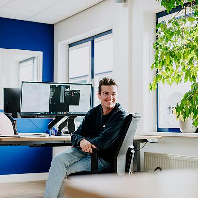 Man zit lachend aan zijn bureau in een modern kantoor met blauwe muren en grote ramen, met meerdere computerschermen en een groene plant naast hem.