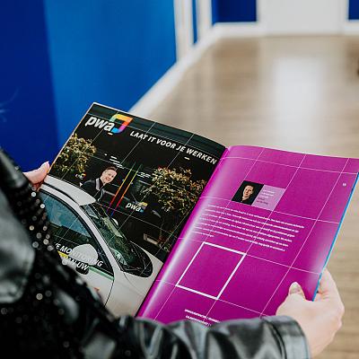 Persoon houdt een open tijdschrift vast met een artikel over PWA, waarbij de linkerpagina een foto toont van een man naast een auto en de rechterpagina tekst bevat op een paarse achtergrond.