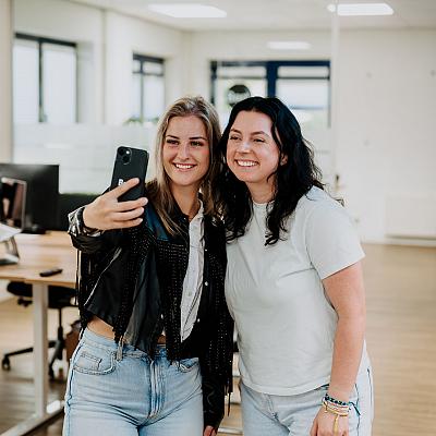 Twee vrouwen maken samen een selfie in een moderne kantoorruimte. Ze lachen en lijken plezier te hebben, terwijl ze voor de camera poseren.