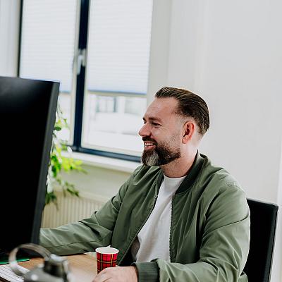 Man zit glimlachend achter zijn bureau in een lichte kantoorruimte, terwijl hij aan het werk is op zijn computer met een koffiebekertje naast zich.