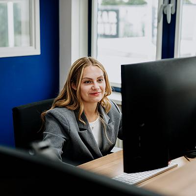 Vrouw met lang blond haar werkt geconcentreerd achter een groot computerscherm in een modern kantoor met blauwe muren.