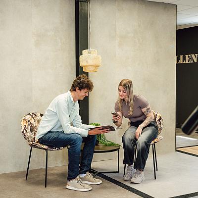 Twee mensen zitten in een moderne showroom op stoelen met bloemenprint en bekijken samen een tegelstaal en een smartphone.