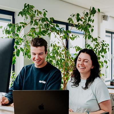 Man en vrouw werken samen achter een laptop in een licht kantoor met veel groen, terwijl ze lachen om iets op het scherm.