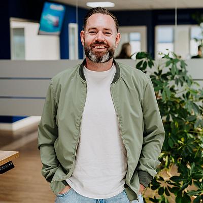 Man met baard glimlacht naar de camera terwijl hij met zijn handen in zijn zakken staat in een modern kantoor met blauwe accenten en groene planten.