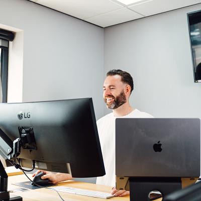Man staat glimlachend achter zijn bureau met twee computerschermen in een modern kantoor met een lichtgrijze muur en een tv-scherm waarop een bedrijfslogo te zien is.