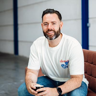 Man zit op een bruine bank in een industriële ruimte, glimlacht naar de camera en houdt een koffiekopje vast.