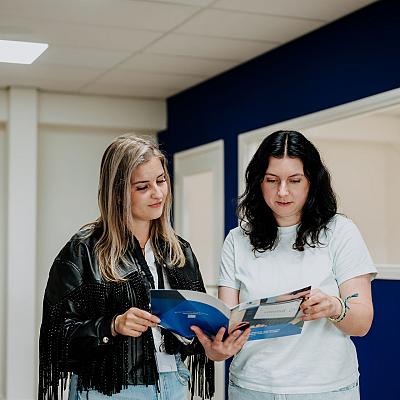 Twee vrouwen staan in een kantoorruimte met blauwe muren en bekijken samen een brochure, terwijl ze in gesprek lijken over de inhoud.