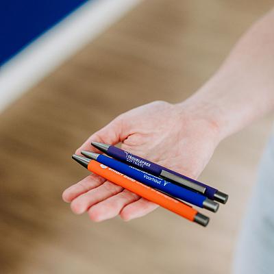 Hand houdt drie pennen vast in de kleuren blauw en oranje met bedrijfslogo’s en teksten erop bedrukt.