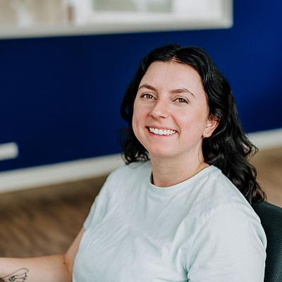 Vrouw met donker haar zit glimlachend aan een bureau in een modern kantoor met blauwe muren.