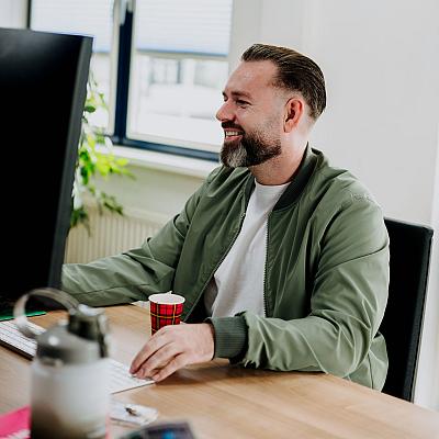 Man zit glimlachend achter zijn bureau in een lichte kantoorruimte, terwijl hij aan het werk is op zijn computer met een koffiebekertje naast zich.