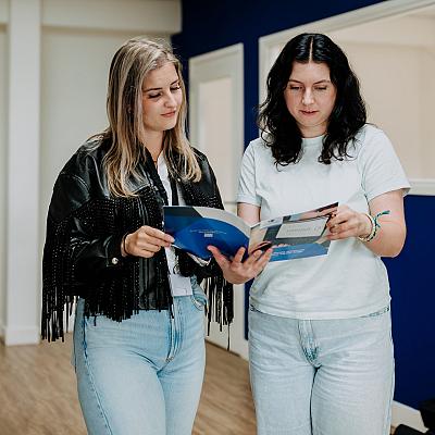 Twee vrouwen bekijken samen een brochure in een moderne kantoorruimte met blauwe muren.