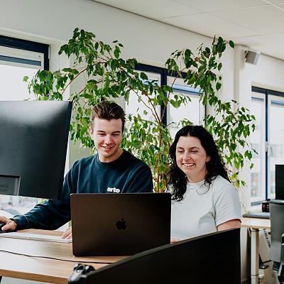 Man en vrouw werken samen achter een bureau in een modern, licht kantoor met grote ramen en groene planten.