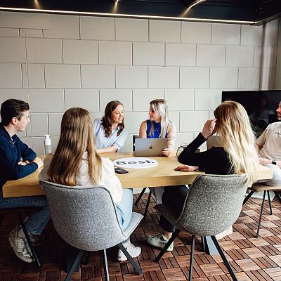 Zes collega’s in gesprek tijdens een teamoverleg aan een houten tafel in een lichte vergaderruimte met een moderne uitstraling.