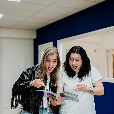 Twee vrouwen kijken enthousiast en verrast naar een open brochure die ze samen vasthouden in een kantoor met blauwe muren.