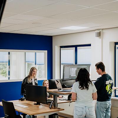Vier collega’s staan en zitten samen rond een bureau in een modern kantoor met blauwe muren en grote ramen, terwijl ze iets bespreken op een computerscherm.