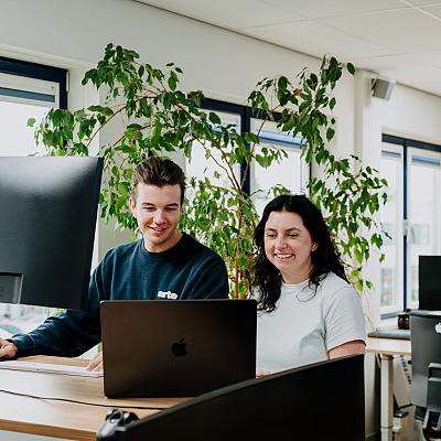 Twee collega’s werken samen achter een bureau in een modern kantoor met veel licht en groene planten.