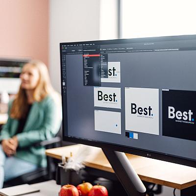 Beeldscherm met het ontwerp van het logo van Best Marketingbureau in Adobe Illustrator, met op de achtergrond een vrouw die op kantoor aan haar bureau zit.