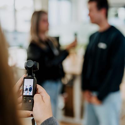 Hand filmt een interview met een handheld camera in een kantooromgeving, terwijl twee collega’s onscherp op de achtergrond met elkaar in gesprek zijn.