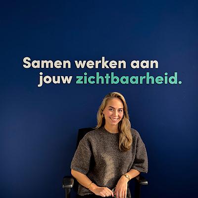 Vrouw met lang blond haar zit op een bureaustoel tegen een donkerblauwe muur, boven haar de tekst “Samen werken aan jouw zichtbaarheid.” waarvan het woord “zichtbaarheid.” in lichtgroen is uitgelicht.