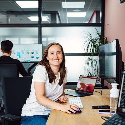 Vrouw die lachend achter haar bureau werkt met laptop en beeldscherm, in een modern kantoor met collega op de achtergrond.