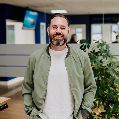 Een glimlachende man met een groene jas, wit T-shirt en lichte spijkerbroek staat met zijn handen in zijn zakken in een kantoor naast een grote plant.