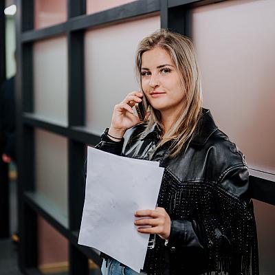 Een vrouw met een zwarte leren jas en een lichte spijkerbroek telefoneert met een vel papier in haar hand, voor een glazen kantoorwand.