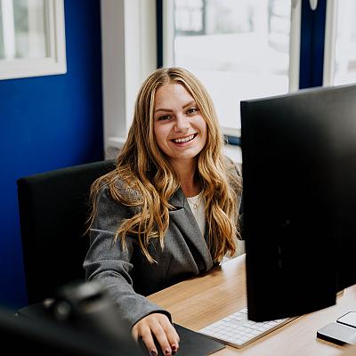 Een glimlachende vrouw met lang blond haar en een donkere blazer zit aan een bureau achter een beeldscherm in een kantoor.
