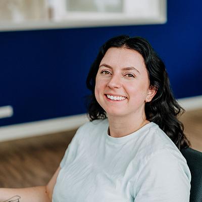 Een glimlachende vrouw met donker haar en een lichtblauw shirt zit op een bureaustoel in een kantoor met een blauwe wand.